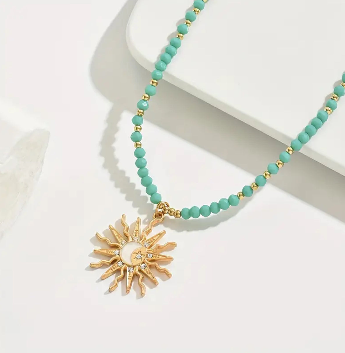 Turquoise beaded necklace with a gold sun pendant on a white surface